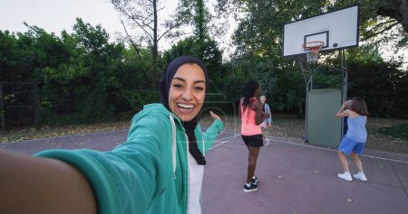 Foto de POV Shot: Grupo de jóvenes adolescentes multiétnicas en ropa deportiva que tienen una videollamada usando un teléfono inteligente. Chicas hablando con un amigo usando Internet en una cancha de baloncesto al aire libre - Imagen libre de derechos