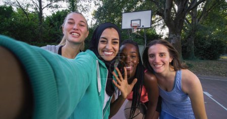 Téléchargez les photos : Portrait d'une amie multiethnique portant des vêtements de sport posant pour un selfie à l'aide d'un téléphone intelligent. Groupe de jeunes femmes célébrant une victoire au terrain de basketball en plein air en prenant une photo pour les médias sociaux - en image libre de droit