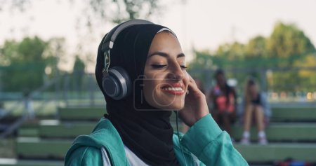 Foto de Primer plano Retrato de una hermosa mujer árabe sonriendo mientras se pone los auriculares en el parque durante su paseo matutino. Adolescente femenina disfrutando de la música y el aire fresco mientras está al aire libre - Imagen libre de derechos