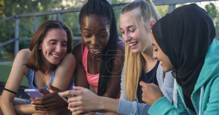 Foto de Grupo de jóvenes adolescentes multiétnicas sentadas en un parque público, riendo y señalando un teléfono inteligente. Grupo de Amigos Revisando sus Memorias y Fotos Infantiles Juntos Usando Tecnología - Imagen libre de derechos