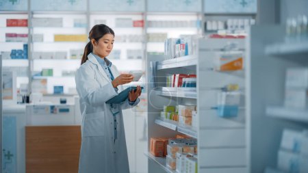 Photo for Pharmacy Drugstore: Beautiful Asian Pharmacist Uses Digital Tablet Computer, Checks Inventory of Medicine, Drugs, Vitamins, Health Care Products on a Shelf. Professional Pharmacist in Pharma Store - Royalty Free Image