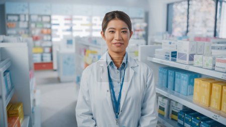 Foto de Farmacia Droguería: Retrato de una hermosa farmacéutica asiática vestida con una capa blanca, mirando a la cámara y sonriendo encantadoramente, detrás de sus estantes llenos de paquetes de medicamentos. Disparo de primer plano medio - Imagen libre de derechos