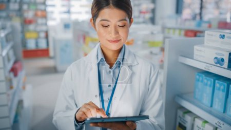 Photo for Pharmacy Drugstore: Portrait of Beautiful Asian Pharmacist Uses Digital Tablet Computer and Smiles Charmingly, Behind Her Shelves Full of Medicine Packages. Medium Close-up Shot - Royalty Free Image