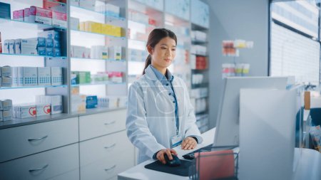 Photo for Pharmacy Drugstore: Beautiful Asian Pharmacist Uses Checkout Counter Computer, Does Inventory Checkup, Online Prescription of Medicine Packages, Drugs, Vitamin Boxes, Supplements, Health Care Products - Royalty Free Image