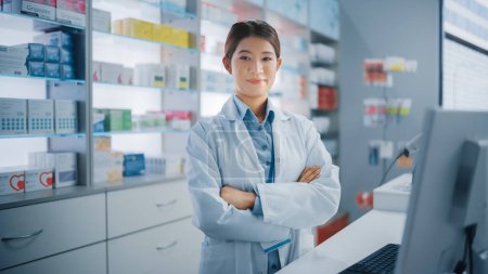 Photo for Pharmacy Drugstore: Portrait of Beautiful Asian Female Pharmacist Wearing White Lab Coat, Looks at Camera, Smiles, Behind Her Checkout Counter, Shelves with Medicine Packages, Health Care Products - Royalty Free Image