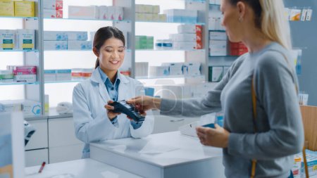 Téléchargez les photos : Pharmacie Drugstore Checkout Caissier Counter : Professional Asian Female Pharmacist Recommande la Médecine dans un Forfait, Blanc Femme Client paie en utilisant le terminal de paiement sans contact et carte de crédit - en image libre de droit