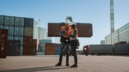Photo for Multiethnic Female Industrial Engineer with Tablet Computer and Male Foreman Worker in Hard Hats and Orange High-Visibility Vests Stand in Container Terminal. Colleagues Talk About Logistics Business. - Royalty Free Image
