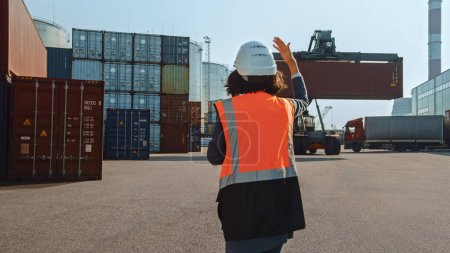 Photo for Female Industrial Engineer with Two-Way Radio Wearing White Hard Hat and Orange High-Visibility Vest Pointing the Unloading Location for the Container Handler in Logistics Operation Terminal. - Royalty Free Image