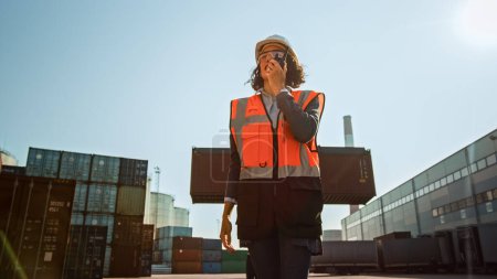 Photo for Female Industrial Engineer with Two-Way Radio Wearing White Hard Hat and Orange High-Visibility Vest Talking with the Container Handler about Unloading Location in Logistics Operation Terminal. - Royalty Free Image