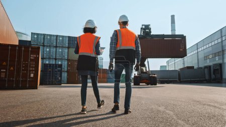 Photo for Multiethnic Female Industrial Engineer with Tablet Computer and Male Foreman Worker in Hard Hats and Orange High-Visibility Vests Walk in Container Terminal. Colleagues Talk About Logistics Business. - Royalty Free Image