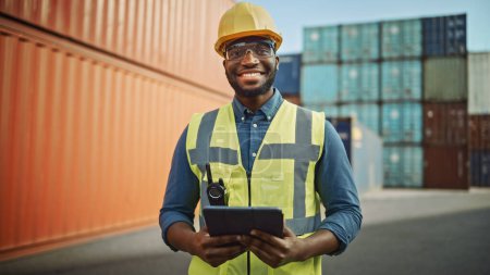 Téléchargez les photos : Portrait souriant d'un bel ingénieur afro-américain noir industriel en casque jaune et gilet de sécurité travaillant sur tablette. Contremaître ou superviseur au terminal à conteneurs. - en image libre de droit