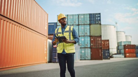 Photo for Smiling Handsome African American Black Industrial Engineer in Yellow Hard Hat and Safety Vest Working on Tablet Computer. Foreman or Supervisor in Container Terminal. - Royalty Free Image