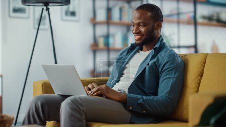 Foto de Hombre negro afroamericano guapo trabajando en la computadora portátil mientras está sentado en un sofá en la acogedora sala de estar. Freelancer Trabajando desde casa. Navegar por Internet, Usar Redes Sociales, Divertirse en Plano. - Imagen libre de derechos