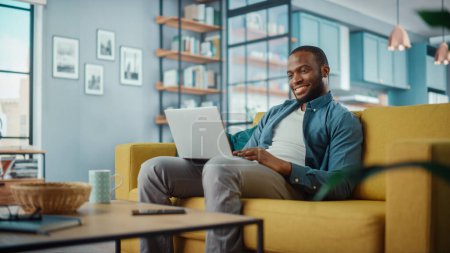 Glücklicher schwarzer Afroamerikaner, der am Laptop arbeitet, während er auf einem Sofa im gemütlichen Wohnzimmer sitzt. Freiberufler, die von zu Hause aus arbeiten. Surfen im Internet, soziale Netzwerke nutzen, Spaß in der Wohnung haben.