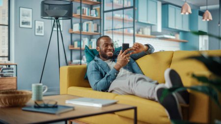 Foto de Emocionado hombre afroamericano negro usando Smartphone mientras descansa en un sofá en la sala de estar. Hombre feliz sonriendo en casa y charlando con colegas y clientes a través de Internet. Usar redes sociales. - Imagen libre de derechos