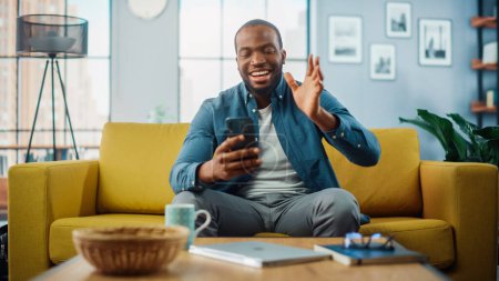 Téléchargez les photos : Happy Black African American Man Avoir un appel vidéo sur Smartphone tout en étant assis sur un canapé dans le salon. Homme excité souriant à la maison et parlant à ses collègues et clients sur Internet. - en image libre de droit