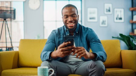 Téléchargez les photos : Homme noir afro-américain excité utilisant un téléphone intelligent tout en se reposant sur un canapé dans le salon. Happy Man Souriant à la maison et bavardant avec ses collègues et ses clients sur Internet. Utiliser les réseaux sociaux. - en image libre de droit