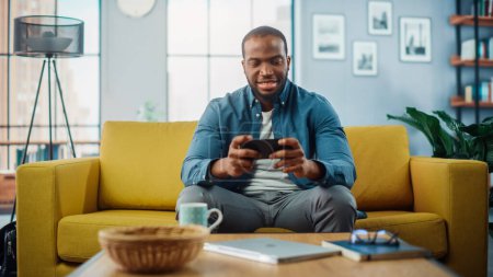 Téléchargez les photos : Heureux homme noir afro-américain ayant un jeu vidéo sur Smartphone App tout en étant assis sur un canapé dans le salon. Personne excitée de couleur reposant à la maison et s'amusant sur l'Internet. - en image libre de droit
