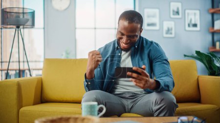 Téléchargez les photos : Heureux noir afro-américain jouer jeu vidéo sur Smartphone App et gagner, montrant OUI geste. Il est assis sur un canapé et se repose dans le salon, s'amusant sur Internet. - en image libre de droit