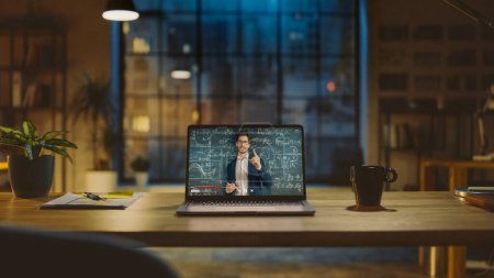 Photo for Shot of a Laptop Computer Standing on the Wooden Desk Showing Online Lecture with Portrait of a Cute Male Teacher Explaining Math Formulas. Modern Apartment with Warm Evening Lighting and Big Window - Royalty Free Image