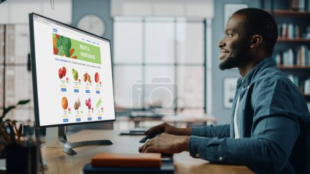Photo for Handsome Black African American is Using Desktop Computer with Groceries Delivery Web Page to Order Fresh Vegetables to Make Dinner. He Lives in Modern Huge Appatment. - Royalty Free Image