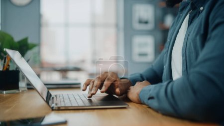 Photo for Close Up on Hands of a Black African American Man Working on Laptop Computer while Sitting Behind Desk in Cozy Living Room. Freelancer Working From Home. Browsing Internet, Using Social Network. - Royalty Free Image