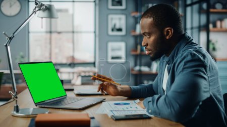 Téléchargez les photos : African American Man Avoir une leçon vidéo en ligne sur ordinateur portable avec écran vert simulé dans le salon. Freelance Travailler sur ses compétences ou étudiant Faire ses devoirs sur Internet. - en image libre de droit
