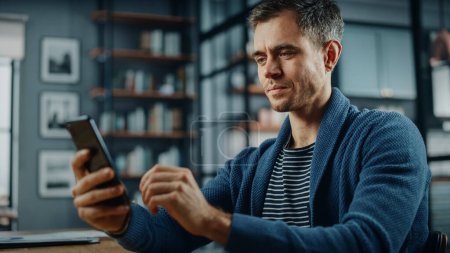 Photo for Serious Caucasian Man Using Smartphone while Sitting at a Desk in Stylish Living Room. Young Man Smiling at Home and Chatting to Colleagues and Clients Over the Internet. Using Social Networks. - Royalty Free Image