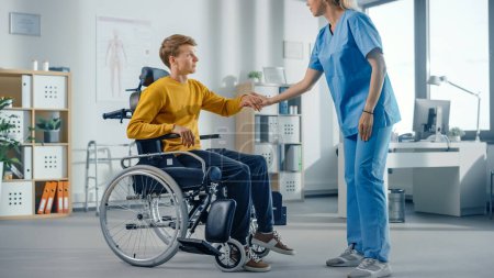 Téléchargez les photos : Physiothérapie hospitalière : un jeune homme atteint d'une blessure déterminé se lève avec succès du fauteuil roulant pour marcher avec l'aide d'un physiothérapeute en réadaptation. Volonté, grain, force d'esprit. - en image libre de droit