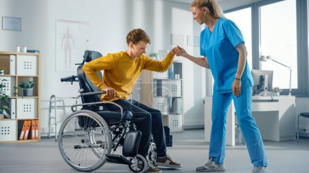 Téléchargez les photos : Physiothérapie hospitalière : Un jeune homme atteint d'une blessure se lève avec succès du fauteuil roulant et marche avec l'aide d'un physiothérapeute en réadaptation. Volonté, grain, force d'esprit. - en image libre de droit