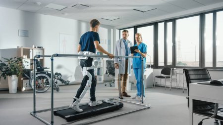 Téléchargez les photos : Physiothérapie hospitalière moderne : Patient avec blessures marche sur tapis roulant portant un exosquelette robotique avancé. Physiothérapie Réadaptation Scientifiques, ingénieurs, médecins utilisent Tablet Computer pour aider - en image libre de droit
