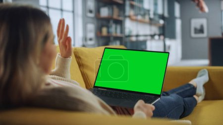 Photo for Beautiful Female Specialist Making a Video Call on Laptop with Green Screen Mock Up Display at Home Living Room while Lying on a Couch Sofa. Freelance Chatting Over the Internet on Social Networks. - Royalty Free Image