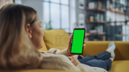 Téléchargez les photos : Belle Authentique Femme Utilisation d'un Smartphone avec écran vert Mock Up Display à la maison Salon tout en s'allongeant sur un canapé. Elle navigue sur Internet et regarde des vidéos sur les réseaux sociaux. - en image libre de droit
