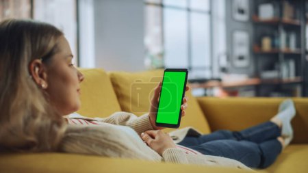 Photo for Beautiful Authentic Female Using a Smartphone with Green Screen Mock Up Display at Home Living Room while Lying on a Couch Sofa. She's Browsing the Internet and Checking Videos on Social Networks. - Royalty Free Image