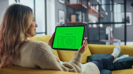 Schöne authentische Frau mit Tablet-Computer mit Green Screen-Mock-Up-Display im heimischen Wohnzimmer, während sie auf einem Sofa liegt. Sie surft im Internet und checkt Videos in sozialen Netzwerken.