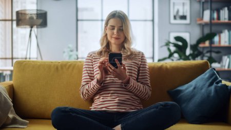 Photo for Beautiful Caucasian Female Using Smartphone in Stylish Living Room while Sitting on a Cozy Couch Sofa. Young Woman Resting at Home, Browsing Internet, Using Social Networks, Having Fun in Flat. - Royalty Free Image