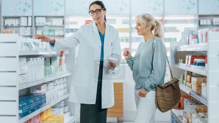Photo for Pharmacy: Professional Specialist Pharmacist Helping Beautiful Senior Female Customer with Medicine Recommendation, Advice, Talking. Drugstore with Full of Drugs, Pills, Health Care, Beauty Products - Royalty Free Image