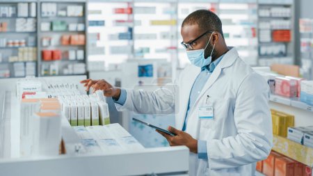 Photo for Pharmacy: Portrait of Professional Black Pharmacist Wearing Face Mask Uses Digital Tablet Computer, Checks Inventory of Medicine, Drugs, Vitamins, Health Care Products on a Shelf. Drugstore Store - Royalty Free Image