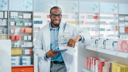 Téléchargez les photos : Pharmacie : Portrait de professionnel confiant pharmacien noir utilise tablette numérique, vérifie l'inventaire de la médecine, regarde la caméra et sourit avec charme. Droguerie avec produits de santé - en image libre de droit