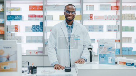 Foto de Pharmacy Drugstore Checkout Counter: Retrato de un guapo profesional negro Hombre Farmacéutico que usa bata blanca de laboratorio, mira a la cámara, sonríe. Estantes con paquetes de medicamentos, productos de cuidado de la salud - Imagen libre de derechos
