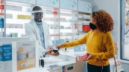 Téléchargez les photos : Pharmacie Drugstore Checkout Counter : Un pharmacien noir professionnel portant un écran facial vend des médicaments à une jeune cliente qui porte un masque facial. Coronavirus Sécurité - en image libre de droit