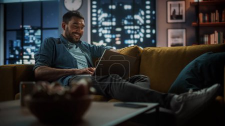 Photo for Beautiful Authentic Latina Female Sitting on Couch Sofa in Cozy Dark Living Room in the Evening and Using Tablet Computer at Home. She's Browsing the Internet, Writing and Drawing with Stylus Pencil. - Royalty Free Image
