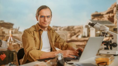 Photo for Archeological Digging Site: Portrait of Great Male Archaeologist Doing Research, Using Laptop, Looks at the Camera Smilingly, Analysing Fossil Remains or Ancient Civilization Culture Artifacts - Royalty Free Image
