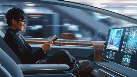 Téléchargez les photos : Concept futuriste : bel homme d'affaires japonais élégant à lunettes lisant un carnet et regardant les nouvelles sur l'écran de réalité augmentée assis dans une voiture autonome à zéro émission. - en image libre de droit