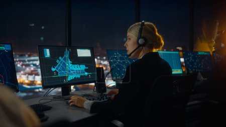 Foto de Controlador de tráfico aéreo femenino con auriculares Hablar en una llamada en la torre del aeropuerto por la noche. La sala de oficina está llena de pantallas de computadora de escritorio con pantallas de navegación, datos de radar de vuelo de avión para el equipo. - Imagen libre de derechos