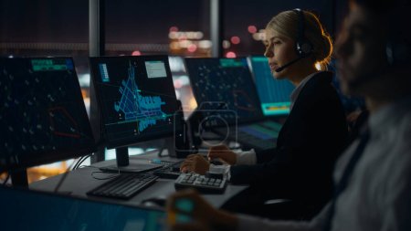 Foto de Controlador de tráfico aéreo femenino con auriculares Hablar en una llamada en la torre del aeropuerto por la noche. La sala de oficina está llena de pantallas de computadora de escritorio con pantallas de navegación, datos de radar de vuelo de avión para el equipo. - Imagen libre de derechos