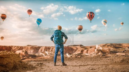 Photo for Young Handsome Tourist Hiking with Backpack in Greater Wilderness in Rocky Canyon Valley. Male Adventurous Backpacker on a Journey Trip. Hot Air Balloon Festival in Mountain National Park. - Royalty Free Image