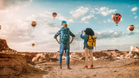 Photo for Young Diverse Tourist Couple Hiking with Backpacks in Great Wilderness in Rocky Canyon Valley. Female Backpacker Taking Photos on Smartphone of Hot Air Balloon Festival in Mountain National Park. - Royalty Free Image