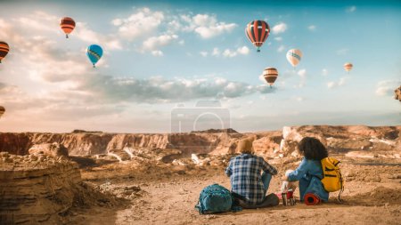 Photo for Young Diverse Tourist Couple Hiking with Backpacks, Resting on Top of Rocky Canyon Valley. Male and Female Backpacker Friends on Adventure Trip. Hot Air Balloon Festival in Mountain National Park. - Royalty Free Image
