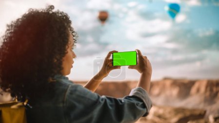 Photo for Excited Young Multiethnic Traveller with Afro Hairstyle Taking a Smartphone Photo on a Phone with Green Screen Display of the Rocky Canyon Valley. Hot Air Balloon Festival in Mountain National Park. - Royalty Free Image